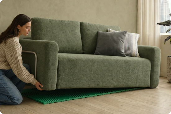 A woman kneels beside a green upholstered sofa in a bright room, touching the front left corner of the sofa. The sofa has two back cushions, two side cushions, and three decorative pillows, including one dark gray pillow and one striped pillow.