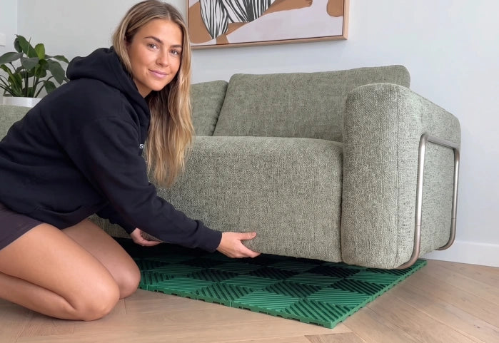 A woman in a black hoodie kneels on a green patterned rug under a light sage green fabric sofa with a chrome metal frame, in a modern room setting.